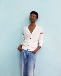 Model wearing a white V-neck cardigan and blue jeans posing against a light blue textured wall.