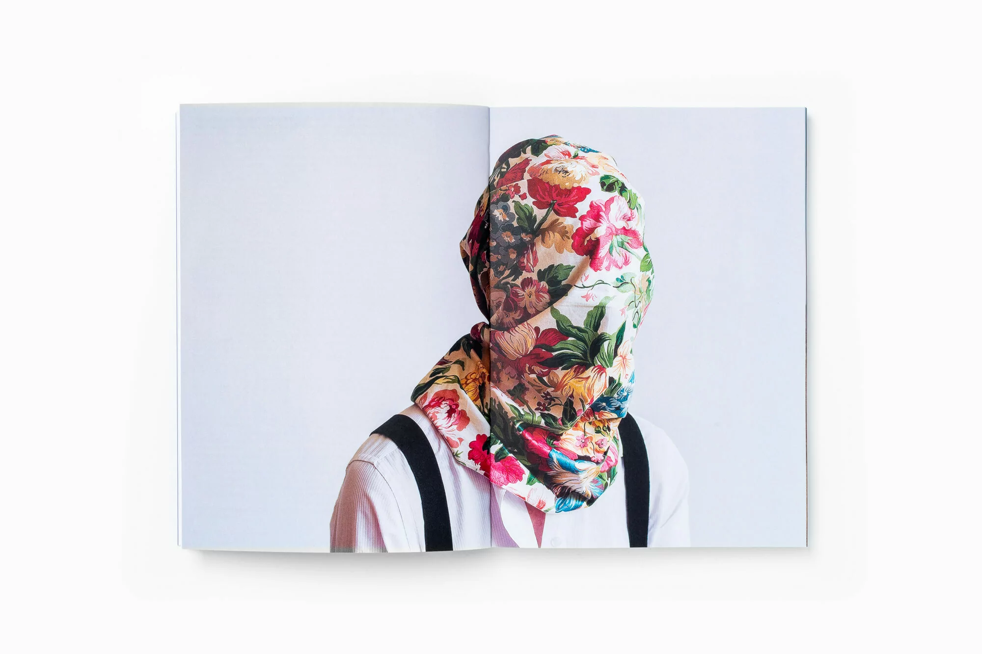 Open book featuring a person with their face covered by a floral patterned scarf, light pink shirt, suspenders, against a white backdrop.