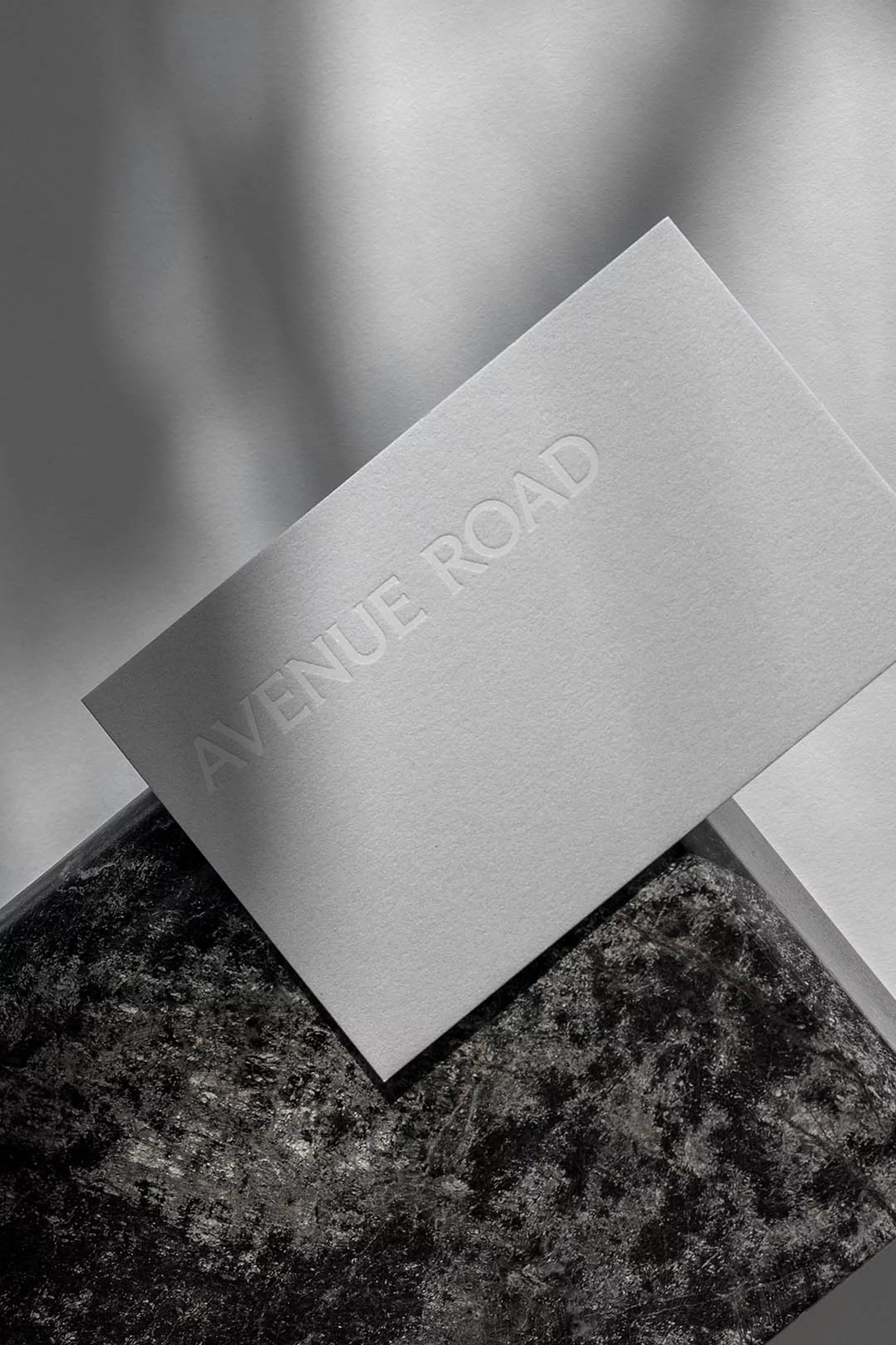 Close-up of a textured white business card with embossed text 'Avenue Road' sitting on a speckled gray stone surface with soft shadows.