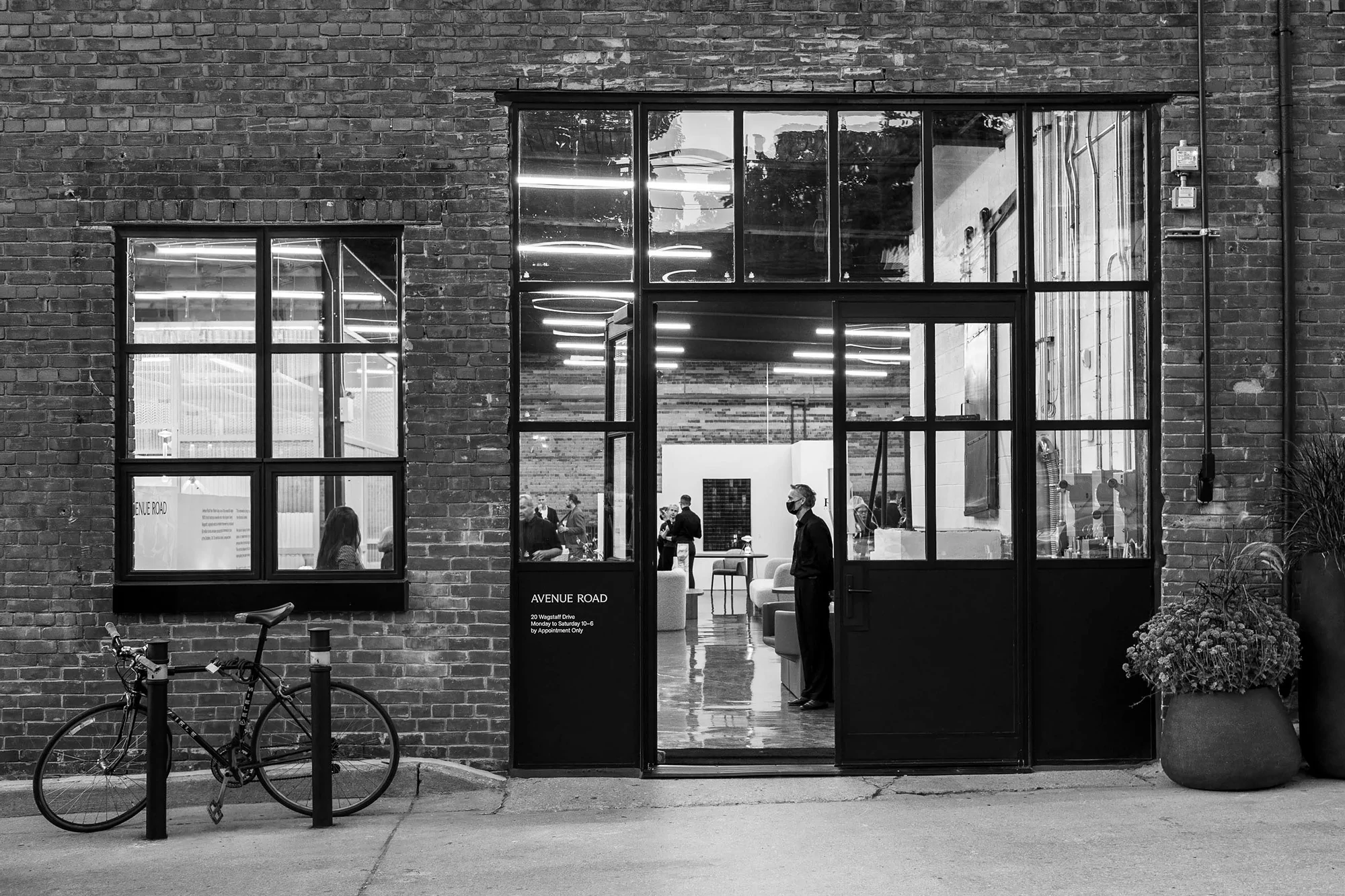 Black and white photo of a bicycle parked outside a brick building with large windows, people inside, and an open glass door.