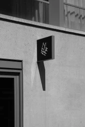 Black and white image of a square branded sign mounted on a textured concrete building exterior wall with harsh sunlight casting a sharp shadow.