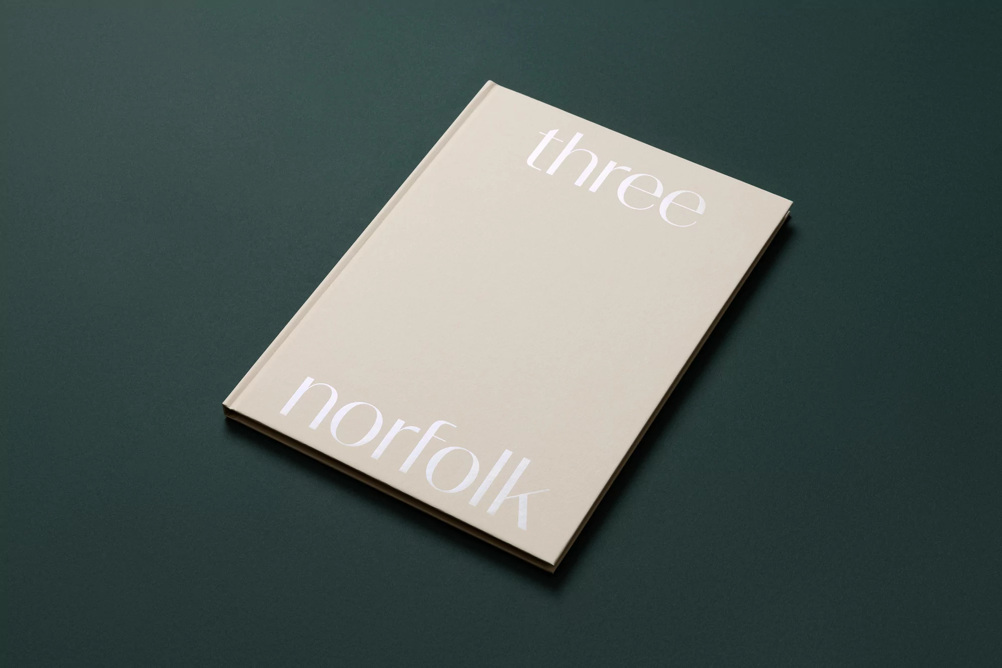 A close-up studio shot features a light beige book with white text, 'three norfolk', placed on a deep green surface.