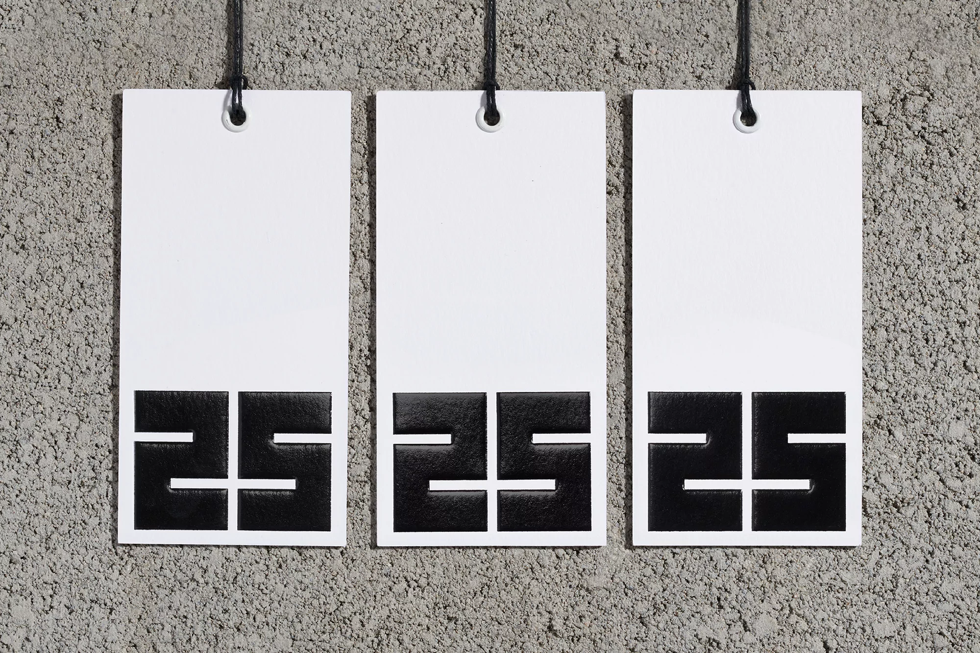 Three white tags hang against a concrete wall, each displaying a bold, black geometric design resembling the number '25'.