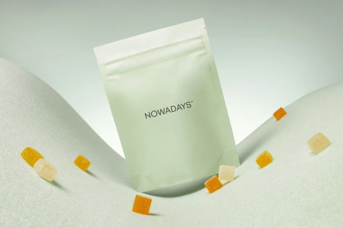 Close-up of a white sealed bag with the text 'NOWADAYS' sitting on a textured white surface with yellow and orange gummies.