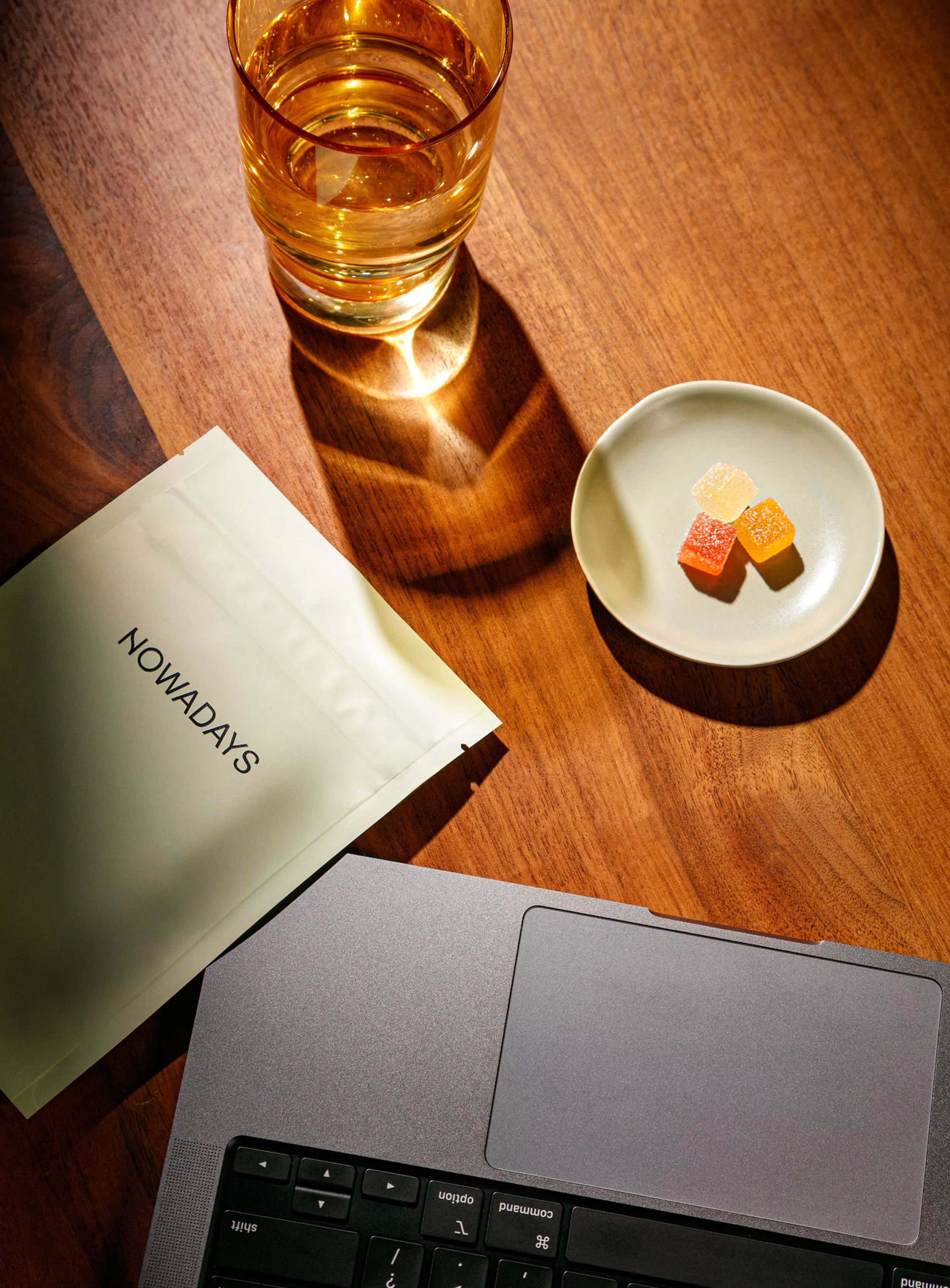 Overhead shot of a laptop, glass of liquid, gummies on a plate, and a sealed package on a wood table in warm, soft lighting.