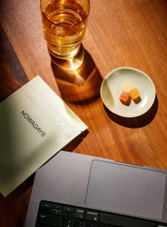 Top-down view of a wooden desk featuring a glass of amber liquid, a small dish with colorful gummies, a pouch, and a laptop keyboard.