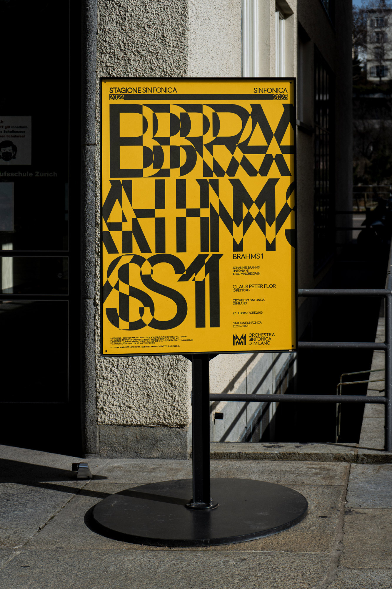 Outdoor poster stand displays concert advertisement with yellow background and bold black typography on stone urban setting.
