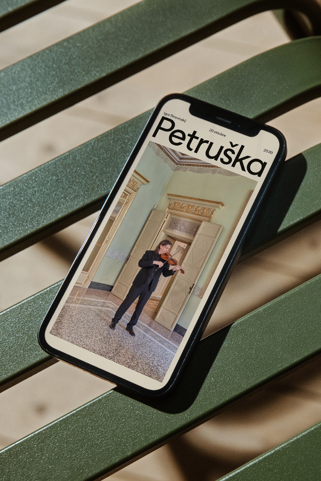 Mobile phone displays 'Petruschka' magazine cover with violinist in ornate room, set against green slatted bench and dappled sunlight.