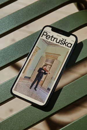 Mobile phone displays 'Petruschka' magazine cover with violinist in ornate room, set against green slatted bench and dappled sunlight.