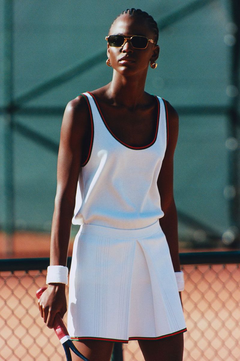 Elegant African-American woman in tennis dress and sunglasses holding racquet on court, in full daylight, showcasing fashion and sport.