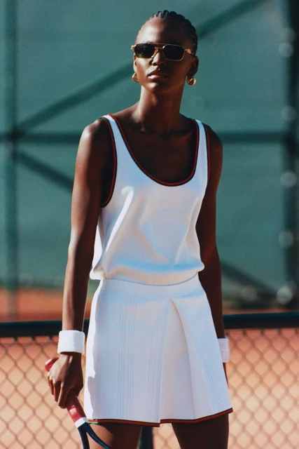 Elegant African-American woman in tennis dress and sunglasses holding racquet on court, in full daylight, showcasing fashion and sport.