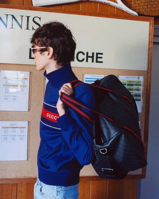 Adult male model with a designer duffel bag stands near a tennis court information board wearing a blue tracksuit jacket and jeans.