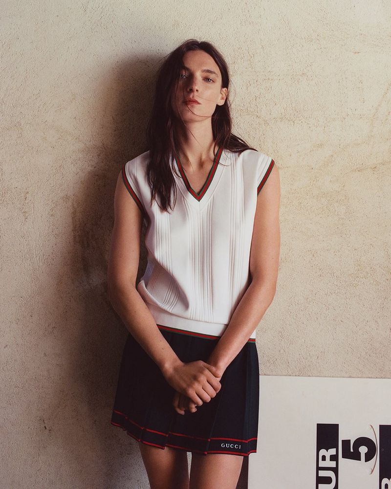 Indoor portrait shot of a woman in a white vest and black skirt leaning against a textured wall with a poster at the bottom.