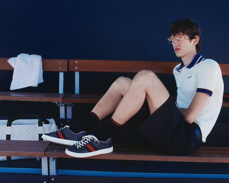 Young man in a polo shirt and shorts sits on a bench next to tennis shoes, a towel, and a Gucci bag in a sportswear ad.