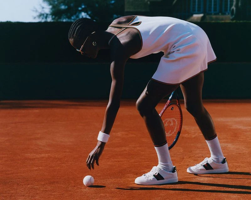 Female tennis player in white dress reaches for ball on red court with racquet visible against dark green background.