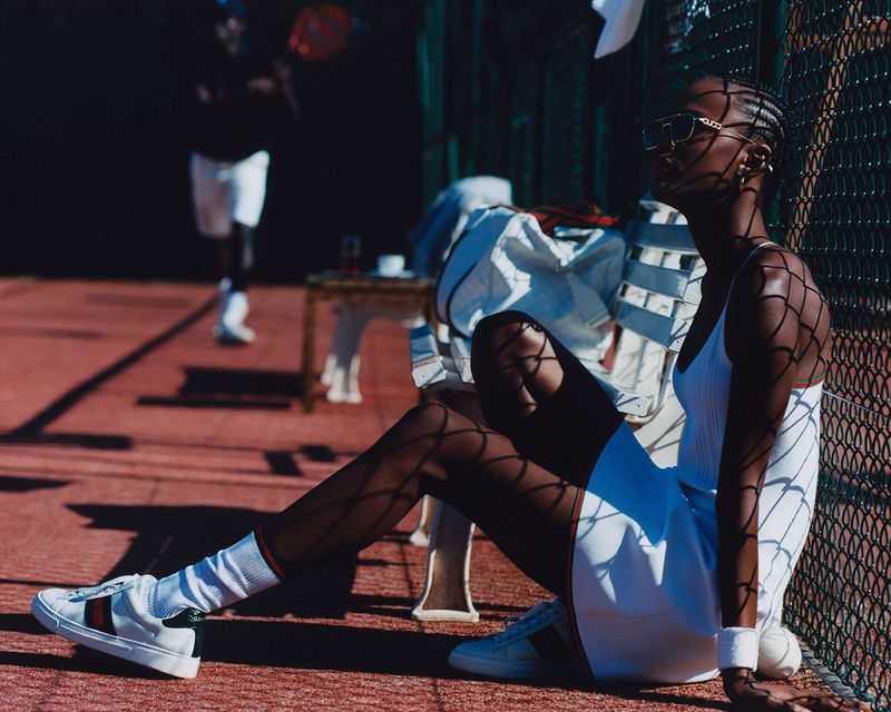 Woman in tennis outfit poses on court, tennis player in background, warm sunny day, low angle, and net shadow.