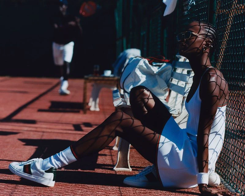 Woman in tennis outfit poses on court, tennis player in background, warm sunny day, low angle, and net shadow.
