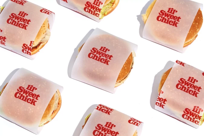 Several 'lil' Sweet Chick' burgers are arranged on a clean white surface, each partially wrapped in branded paper with a visible burger.