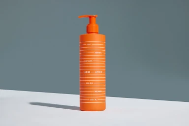 Bright orange cylindrical bottle with a pump dispenser standing on a white surface against a plain grey background.
