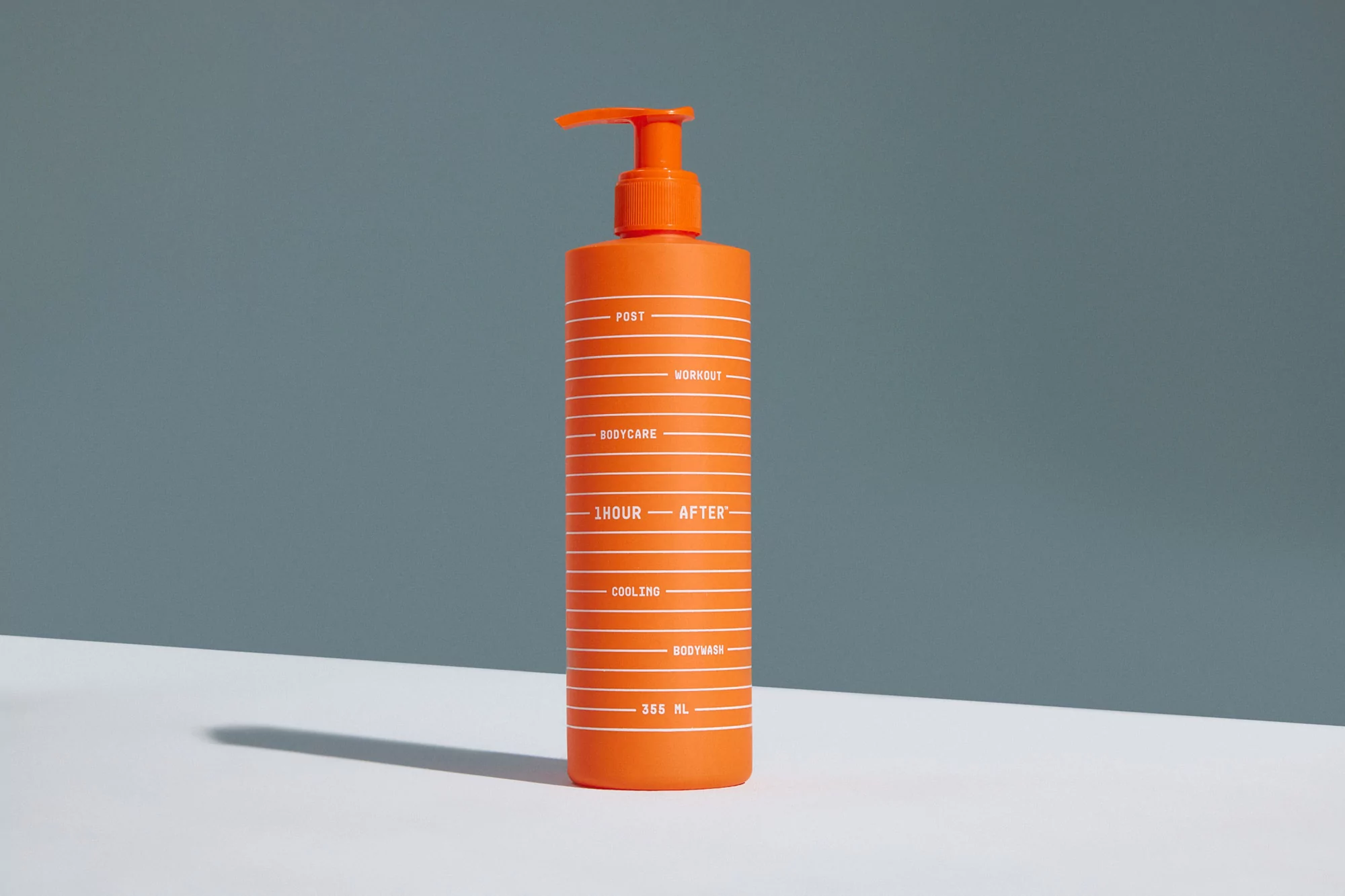 Orange bodywash bottle with pump dispenser sitting on a white surface against a gray backdrop in a brightly lit, neutral product shot.