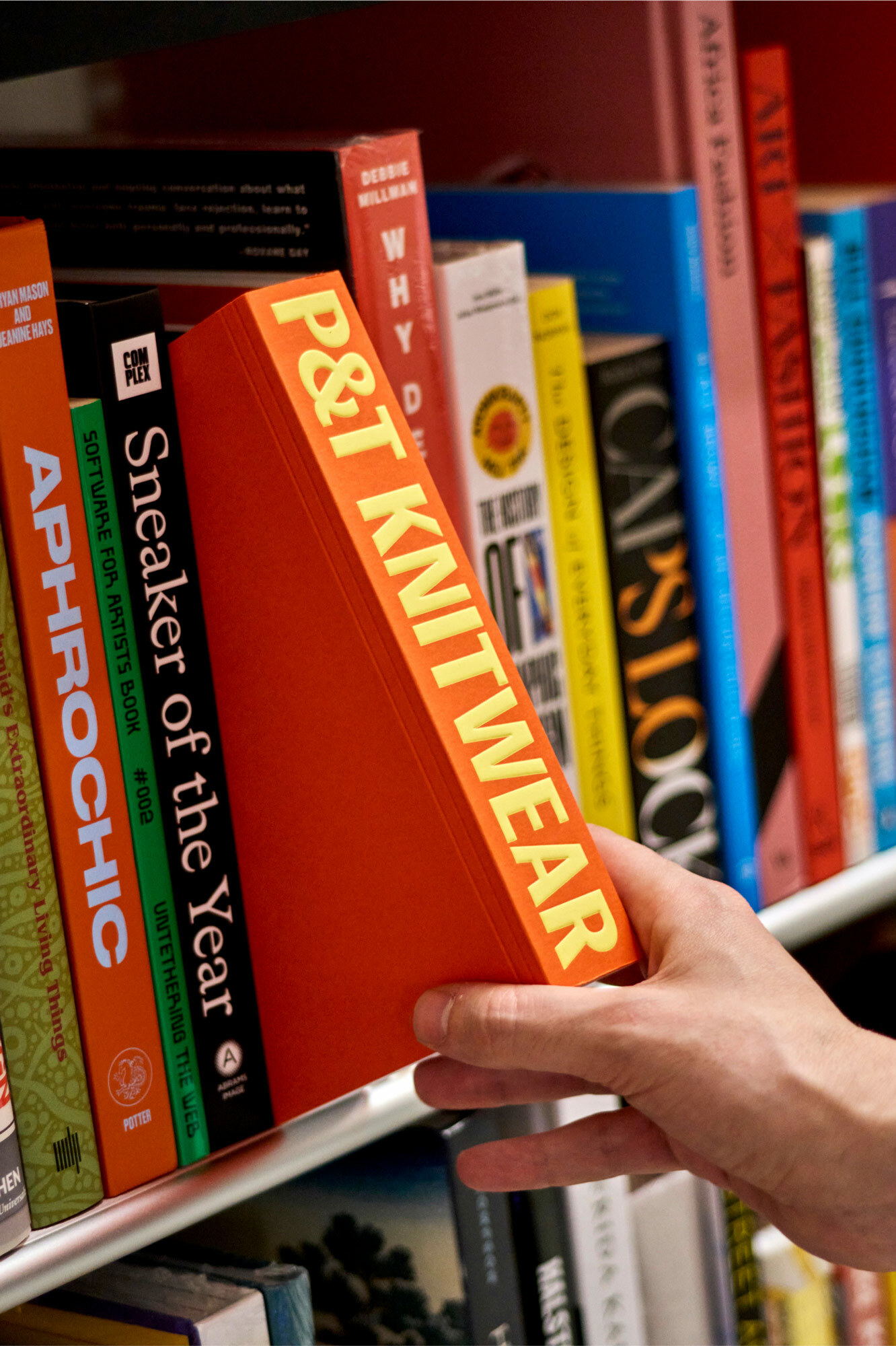 Close-up shot of a hand taking an orange book titled P&T Knitwear from a shelf full of books, creating a library or bookstore scene.