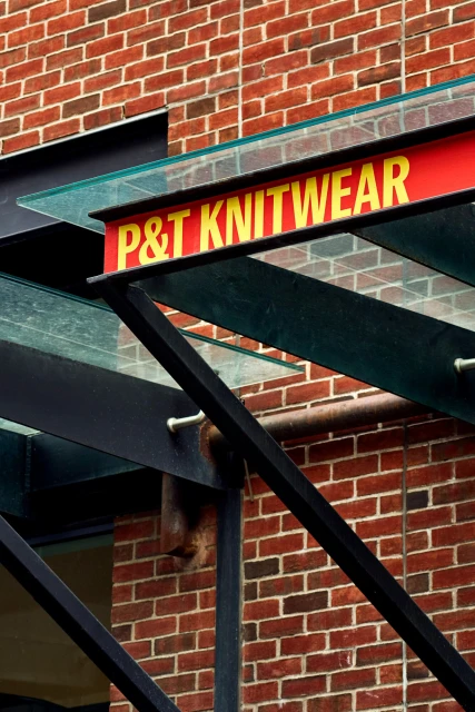 Building exterior featuring a brick wall, a black metal structure, and a red sign with yellow text reading P & T Knitwear.