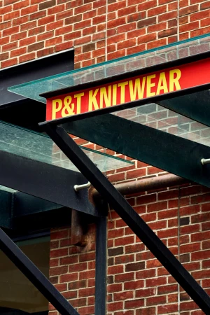 Building exterior featuring a brick wall, a black metal structure, and a red sign with yellow text reading P & T Knitwear.