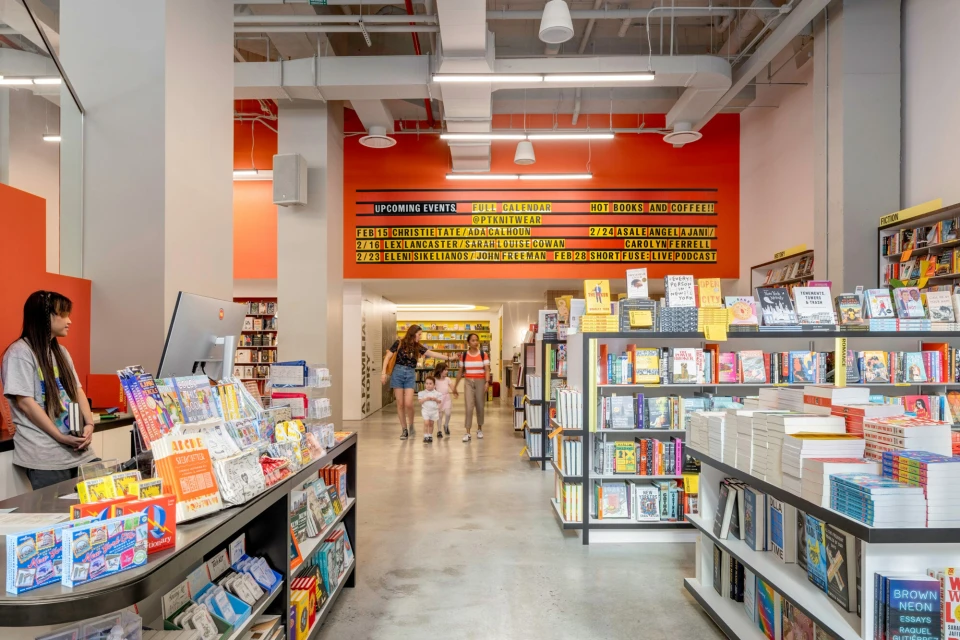 Interior view of a bookstore with shelves stocked with books, people browsing, and a sign listing upcoming events on a bright orange wall.