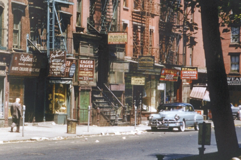Vintage street scene in New York City with classic car parked in front of shops, pedestrians on sidewalk, and buildings with fire escapes.