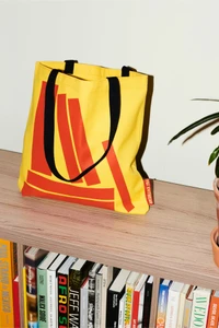Yellow canvas tote bag with bold red geometric graphics and black handles resting on a wooden bookshelf shelf.