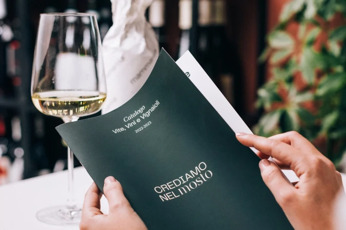 Person holds a catalog with wine in the background. Catalog displays text, 'Crediamo nel Mosto', next to a wine glass and bottle.