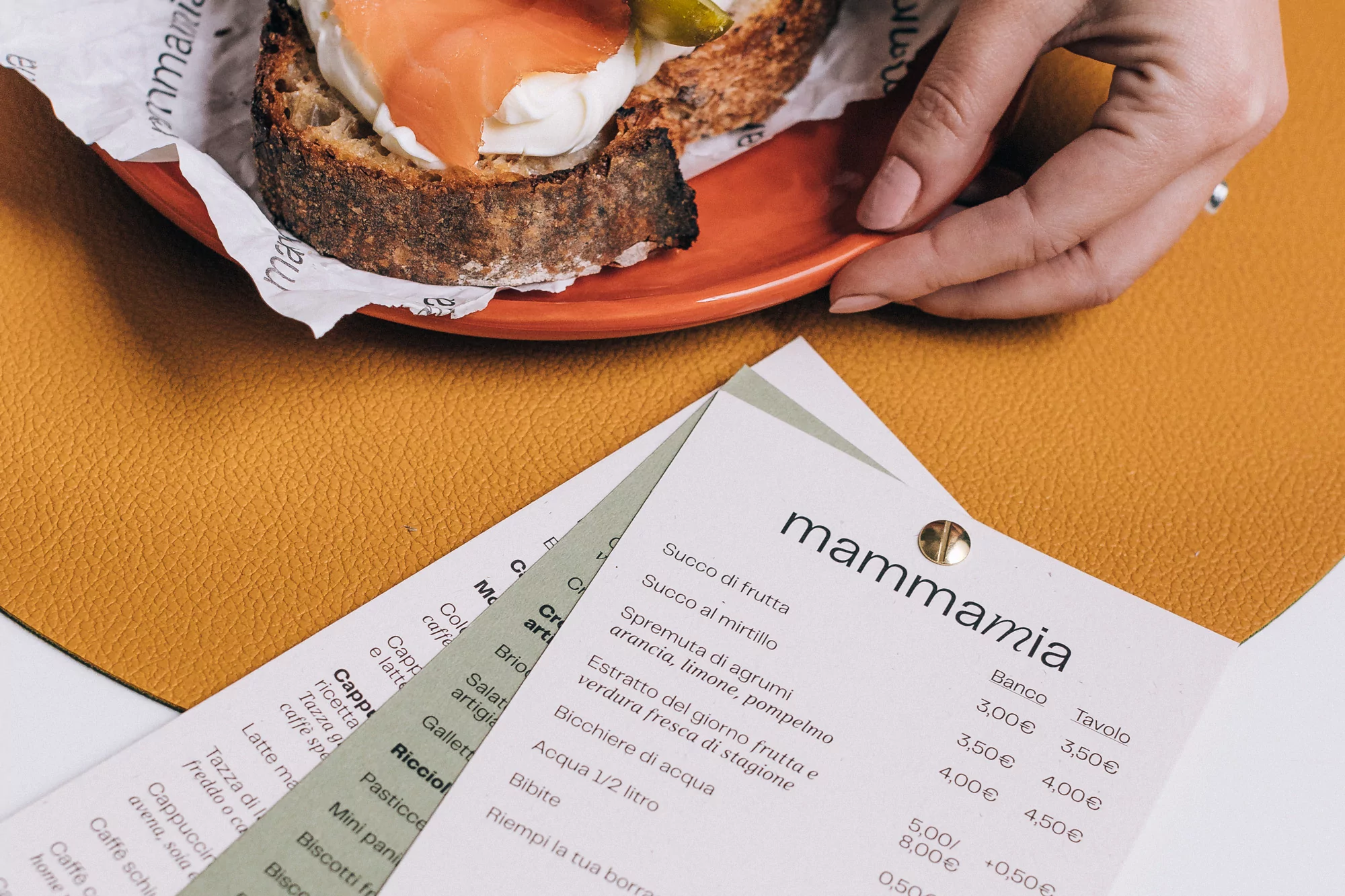 Salmon toast with cream cheese on an orange plate held by a woman's hand, with a restaurant menu on a yellow surface.