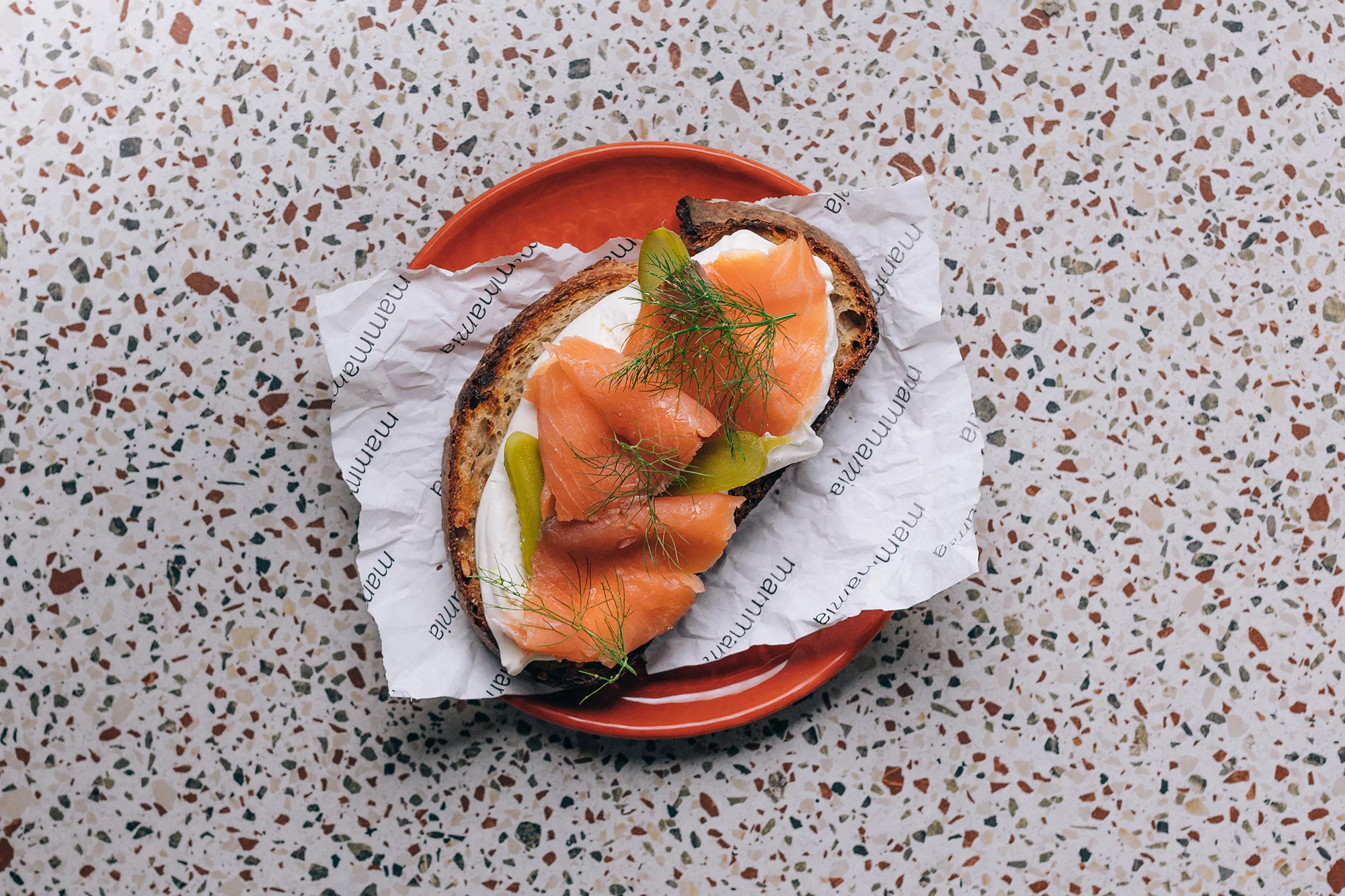 Overhead shot of smoked salmon toast with cream cheese, pickles, and dill sprigs served on an orange plate with a terrazzo background.