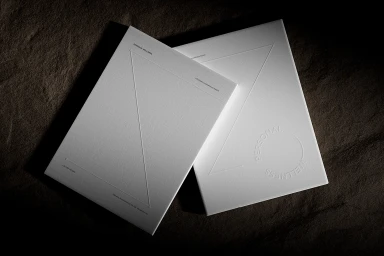Two white embossed stationery cards resting on a dark textured stone surface under dramatic side lighting.