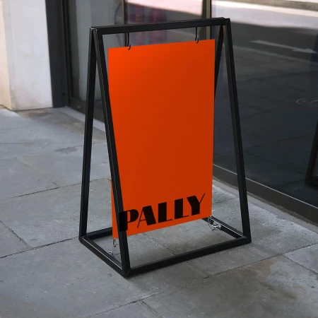 Orange metal A-frame sign stands on a sidewalk with black frame and text 'PALLY'.