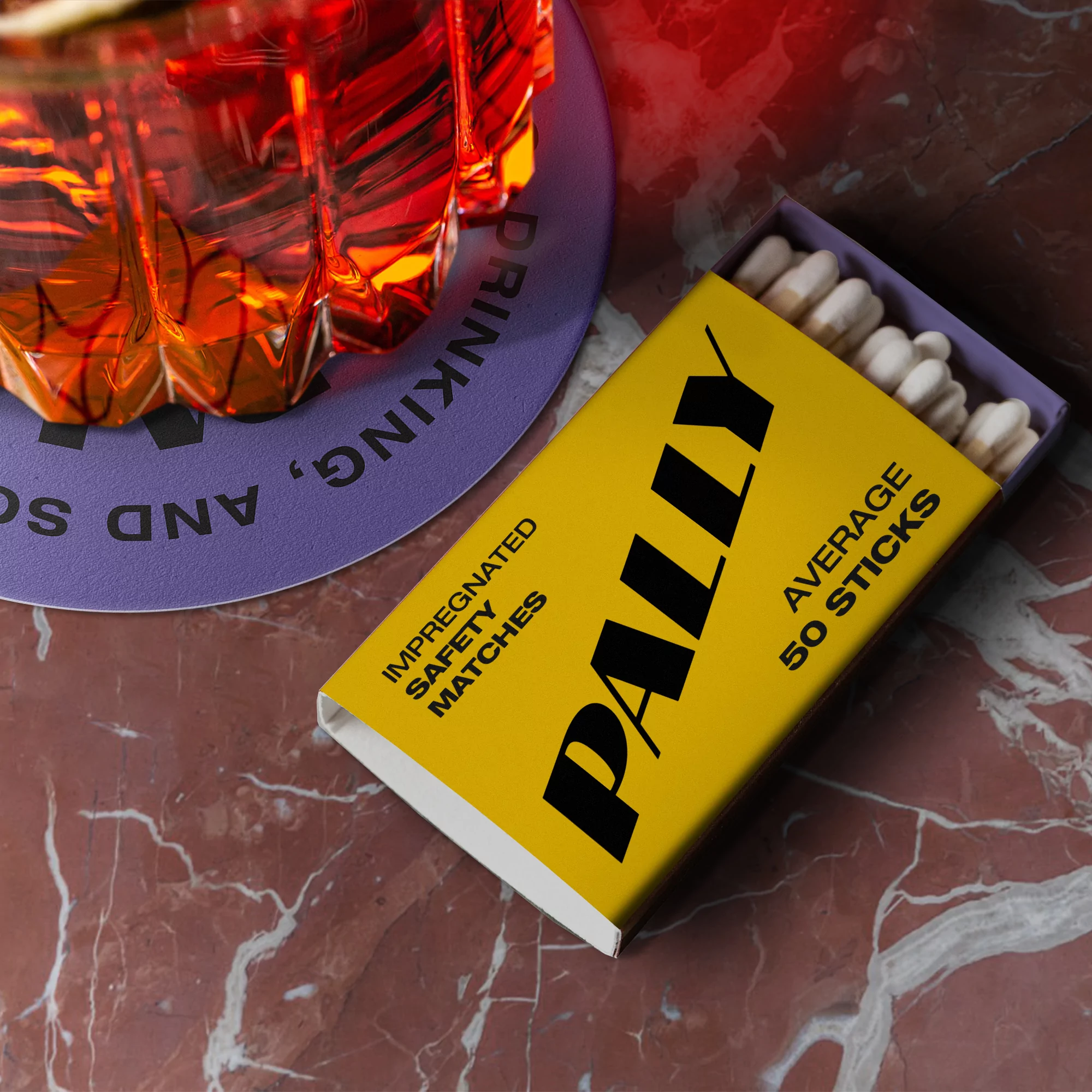 Close-up of an open yellow matchbox with white matches, a partially visible cocktail, and a round purple coaster on a marble surface.