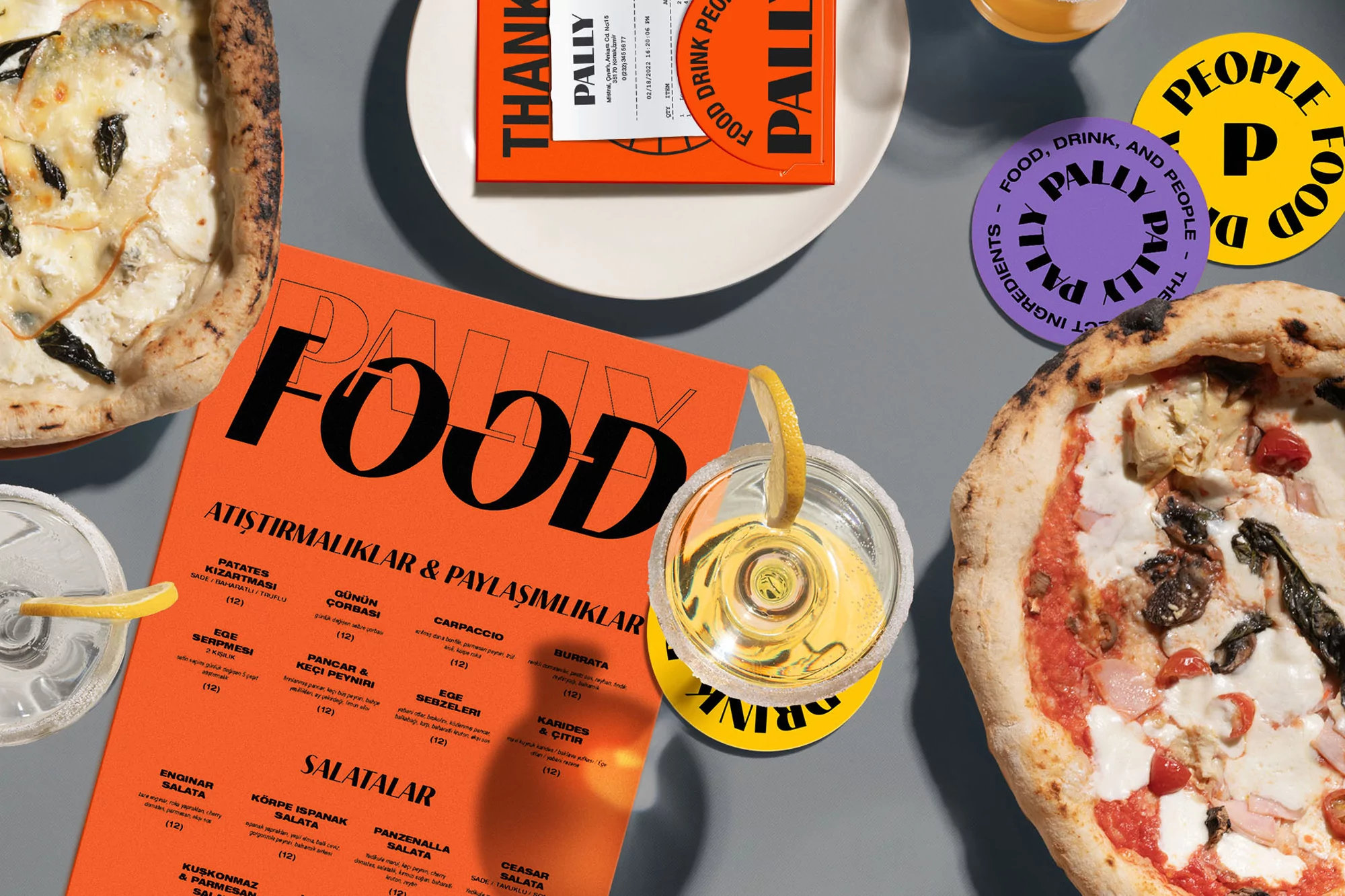 Overhead shot of two pizzas, a clear cocktail with a lemon slice, and bright orange Pally Food restaurant materials on a grey surface.