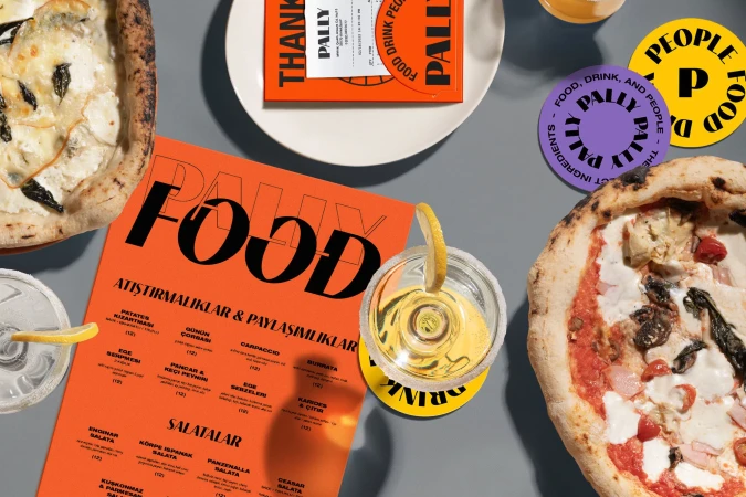 Overhead shot of two pizzas, a clear cocktail with a lemon slice, and bright orange Pally Food restaurant materials on a grey surface.