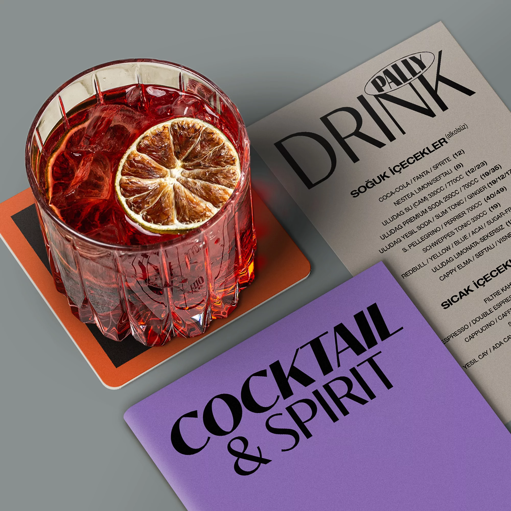A red cocktail drink in a textured glass on a coaster, accompanied by menus, features a slice of dried citrus as garnish.