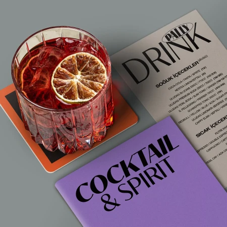 A red cocktail drink in a textured glass on a coaster, accompanied by menus, features a slice of dried citrus as garnish.