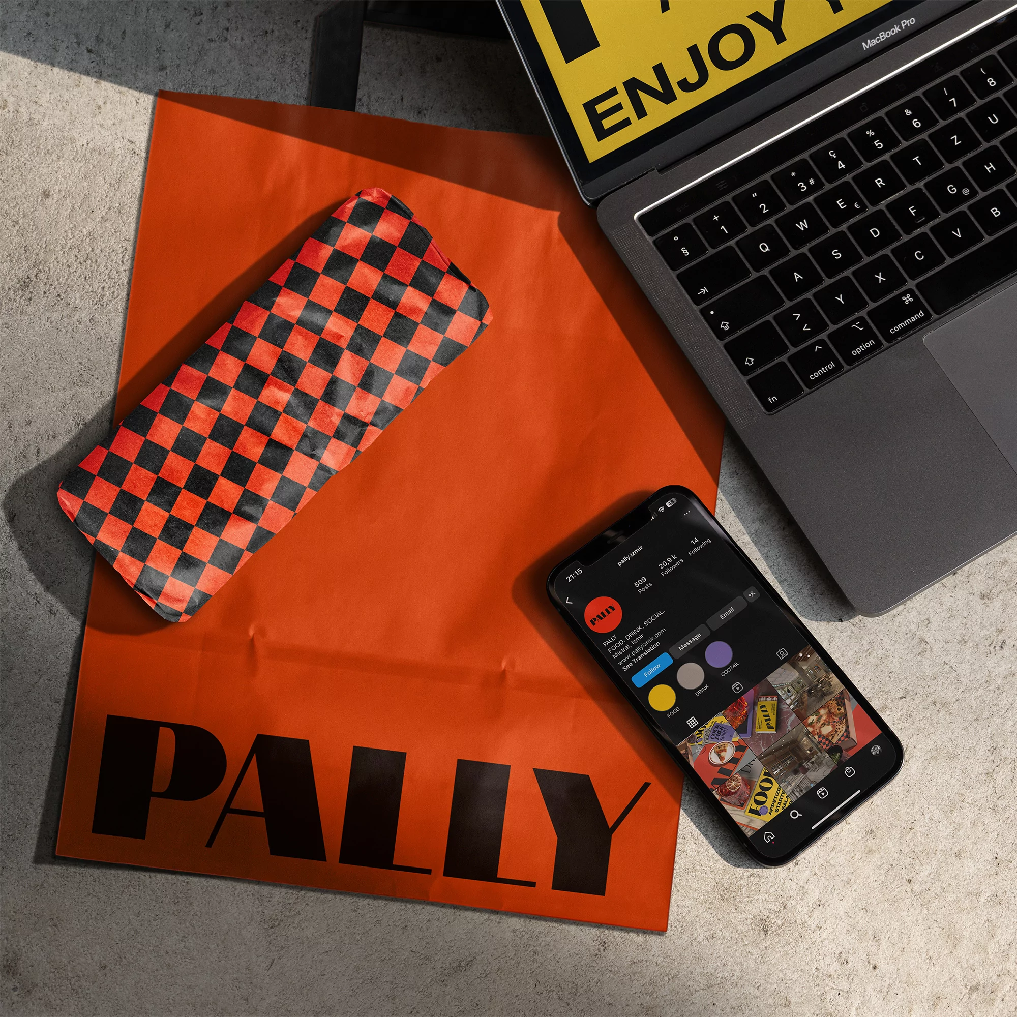 High-angle shot of a laptop, smartphone displaying social media, orange shopping bag, and checkered glasses case on a concrete surface.