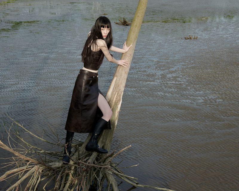 A solitary young woman with dark hair strikes a pose on a fallen tree branch in a marshy, open landscape, wearing a black top, skirt, and boots.
