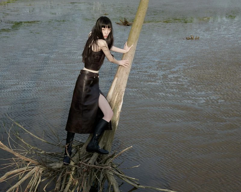 A solitary young woman with dark hair strikes a pose on a fallen tree branch in a marshy, open landscape, wearing a black top, skirt, and boots.