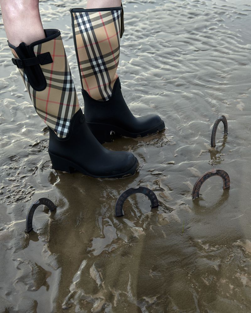 Person wearing black Burberry plaid rain boots stands in wet sand with several rusted metal horseshoes partially buried in the sand.