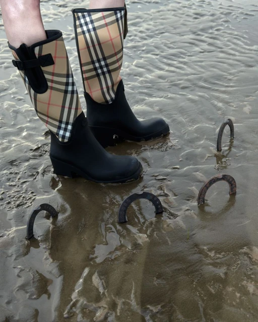 Person wearing black Burberry plaid rain boots stands in wet sand with several rusted metal horseshoes partially buried in the sand.