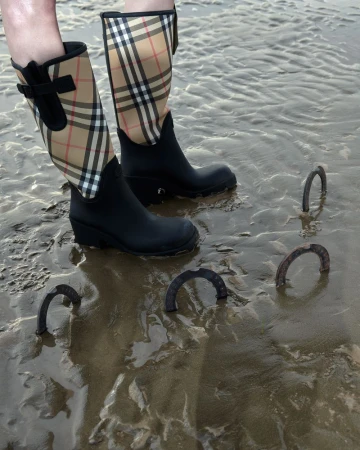 Person wearing black Burberry plaid rain boots stands in wet sand with several rusted metal horseshoes partially buried in the sand.