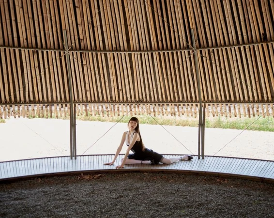 A brunette woman in a black dress and ballet flats is seated on a wooden floor beneath a domed bamboo structure, posing outdoors in natural light.