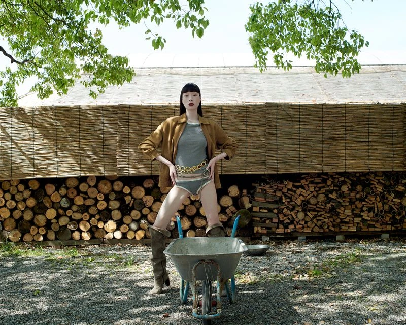 Young woman posing with a wheelbarrow on a gravel road near a wood pile, wearing a light brown jacket and boots in an outdoor fashion shot.