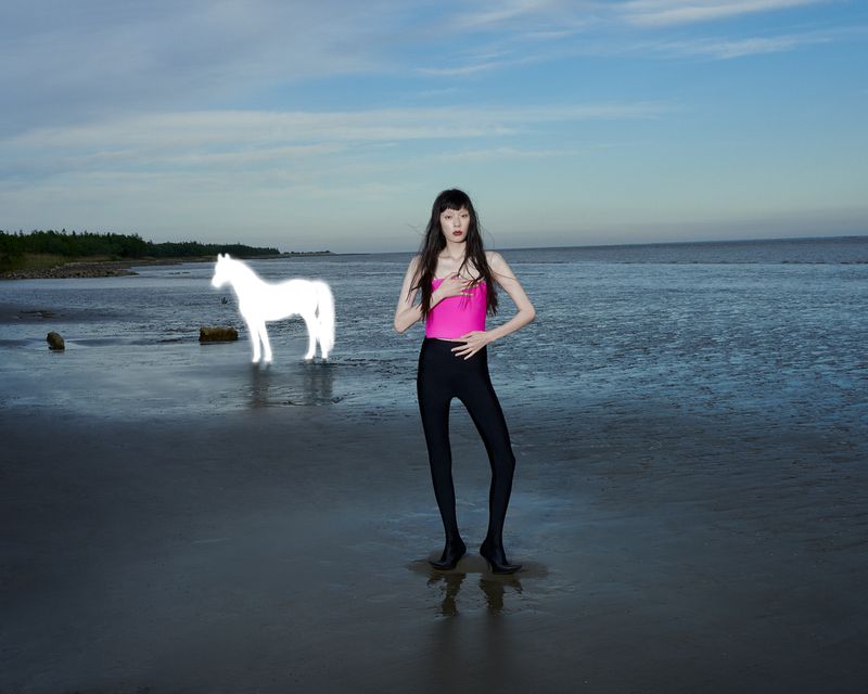 A slender woman poses on a beach near a glowing horse apparition, wearing a pink top and black leggings, with the ocean and sky visible.
