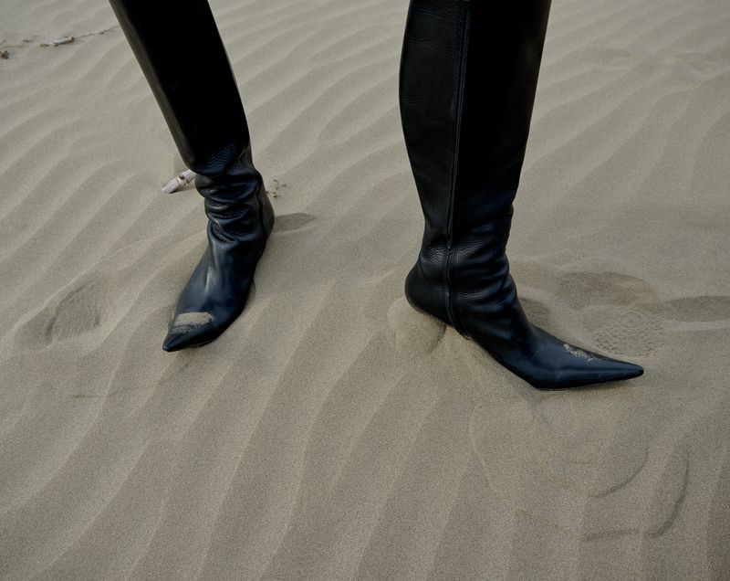 Close-up of a person wearing sleek, black leather knee-high boots standing on a textured sand dune, creating a fashion-forward desert aesthetic.